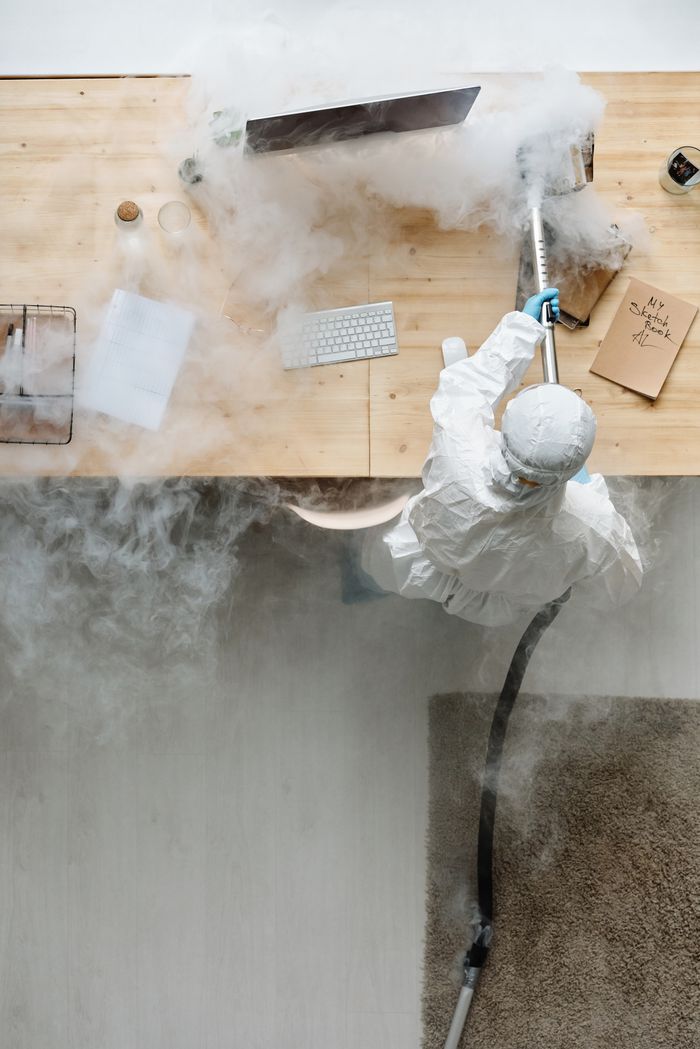 project-02-d A person in protective gear sanitizing an office desk using fumigation equipment during daytime.